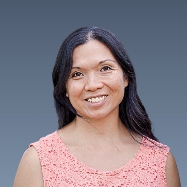 Smiling woman with long dark hair wearing a pink lace top against a gray background.