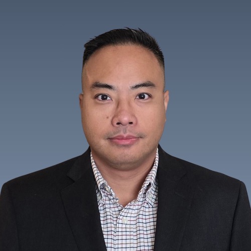 Asian man with short dark hair wearing a dark blazer and patterned shirt against a blue-gray background.