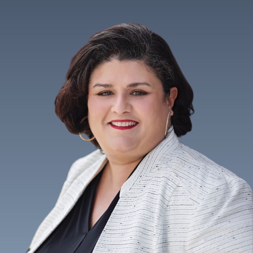 Smiling woman with dark curly hair wearing white blazer and red lipstick against blue background.