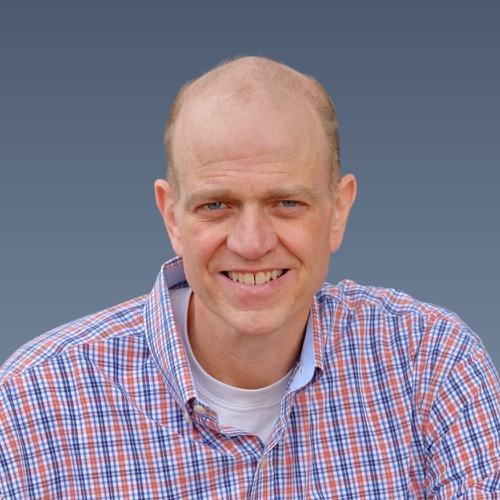 Smiling bald man wearing plaid button-up shirt over white t-shirt against blue-gray background.