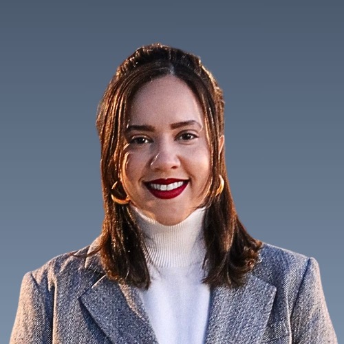 Woman with brown hair, red lipstick, wearing white turtleneck and gray coat against blue background.