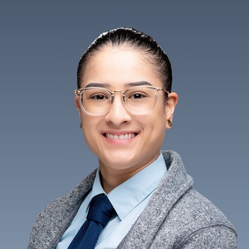 Smiling person with glasses, slicked-back hair, blue shirt, navy tie, and gray jacket against blue background.