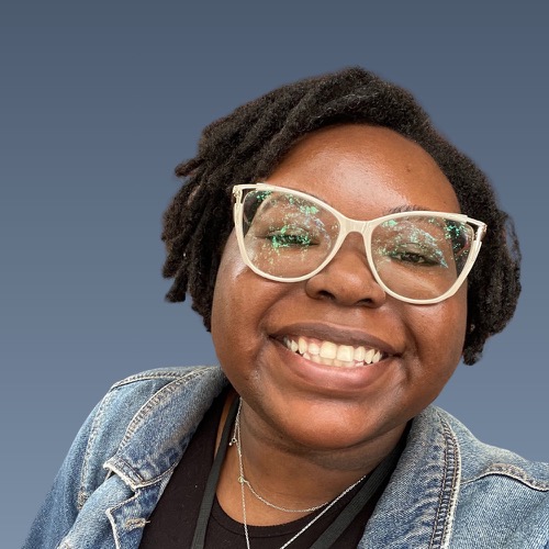 Smiling person with short dreadlocks, white-framed glasses, wearing denim jacket against blue background.