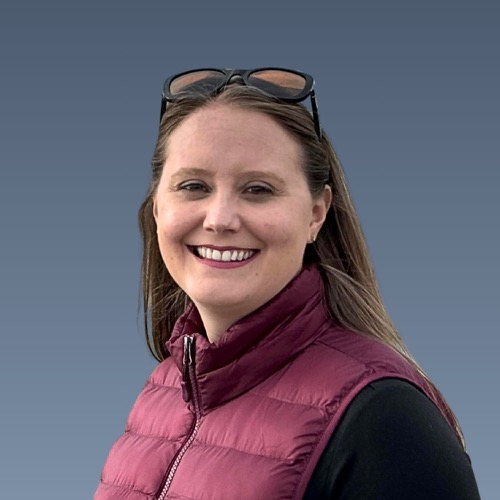 Smiling woman with long hair wearing sunglasses on head and burgundy vest against blue sky background.