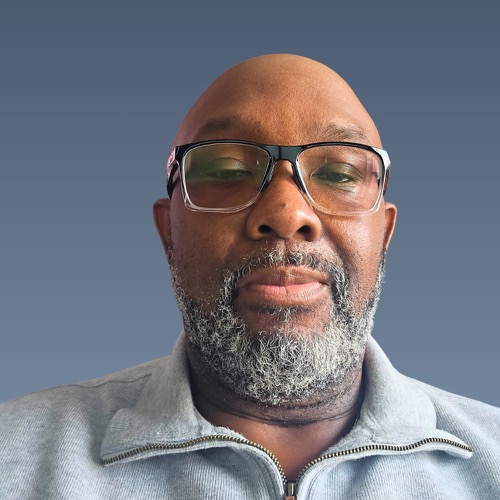 Close-up portrait of a bald man with glasses and a gray beard wearing a light-colored zip-up jacket against a blue background.