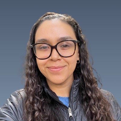 Smiling woman with long curly hair, black-framed glasses, wearing dark jacket against blue-gray background.
