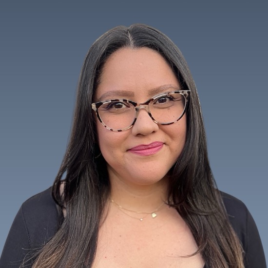 Woman with long dark hair, patterned glasses, pink lips, wearing dark top against blue-gray background.