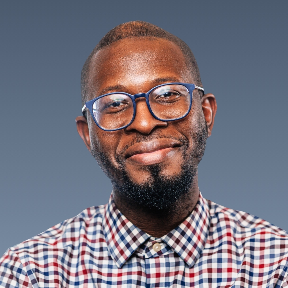 Smiling man with beard, blue glasses, wearing plaid shirt against blue background.