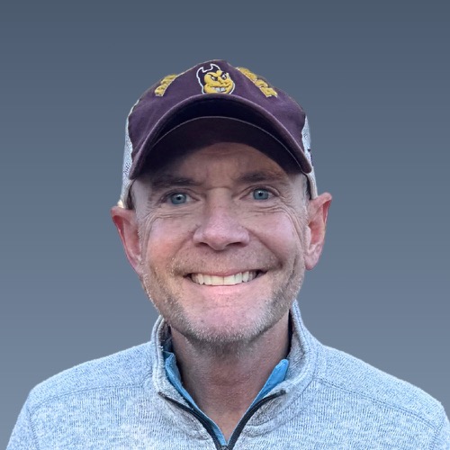 Smiling older man with stubble wearing purple baseball cap and gray sweater against blue background.
