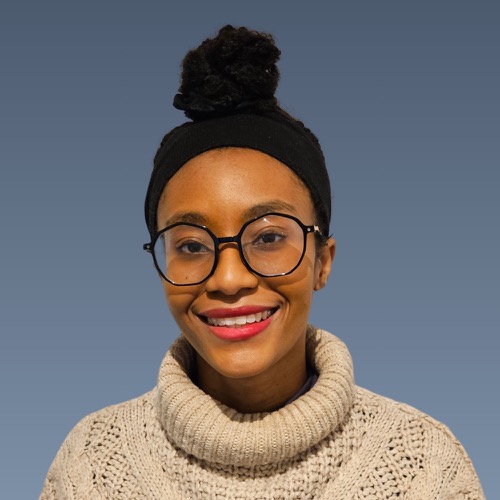 Smiling Black woman with hair in bun, glasses, red lipstick, wearing cream sweater against blue background.