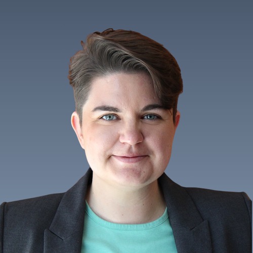 Person with short brown hair and blue eyes wearing black blazer over mint green shirt against blue background.